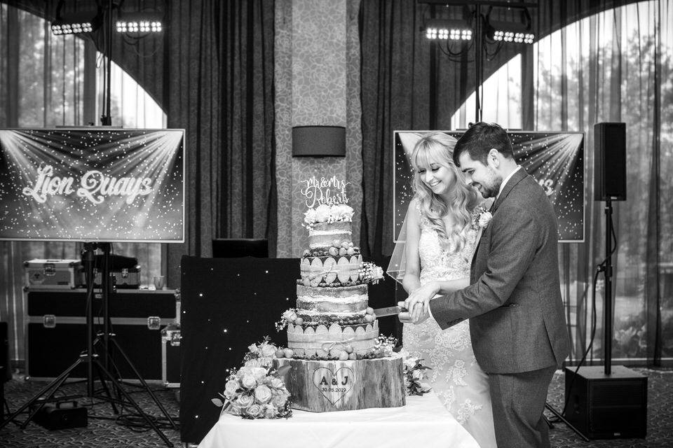 Bride & groom cut wedding cake in front of televisions saying Lion Quays