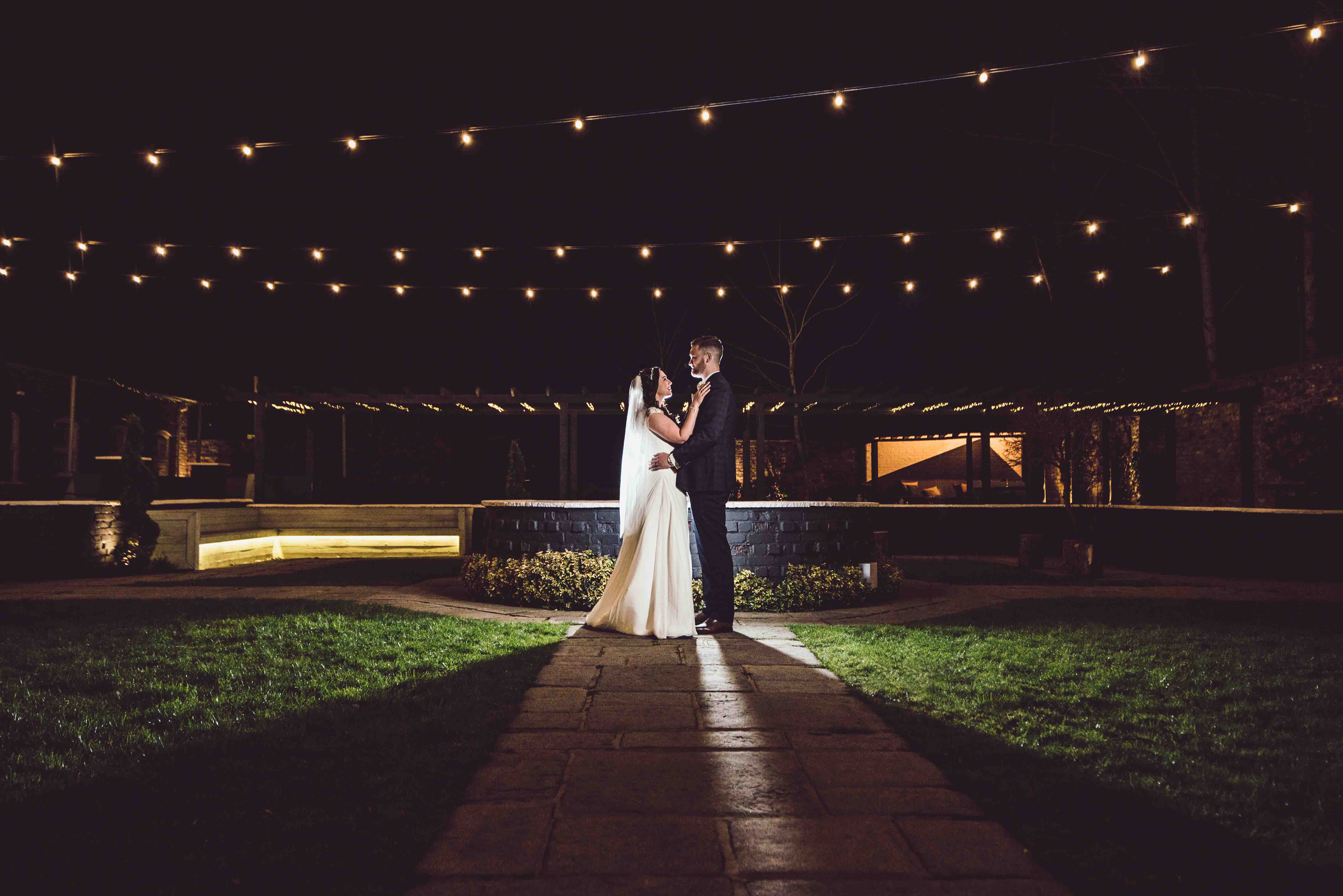 Bride & Groom embrace outside Lion Quays wedding venue at night time with fairy lights above