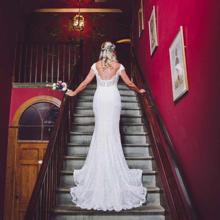 Bride stands on stairs in wedding dress at Harrison's Hall wedding venue, Flintshire