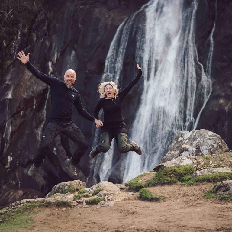 Couple jump with arms out in front of Aber Falls North Wales
