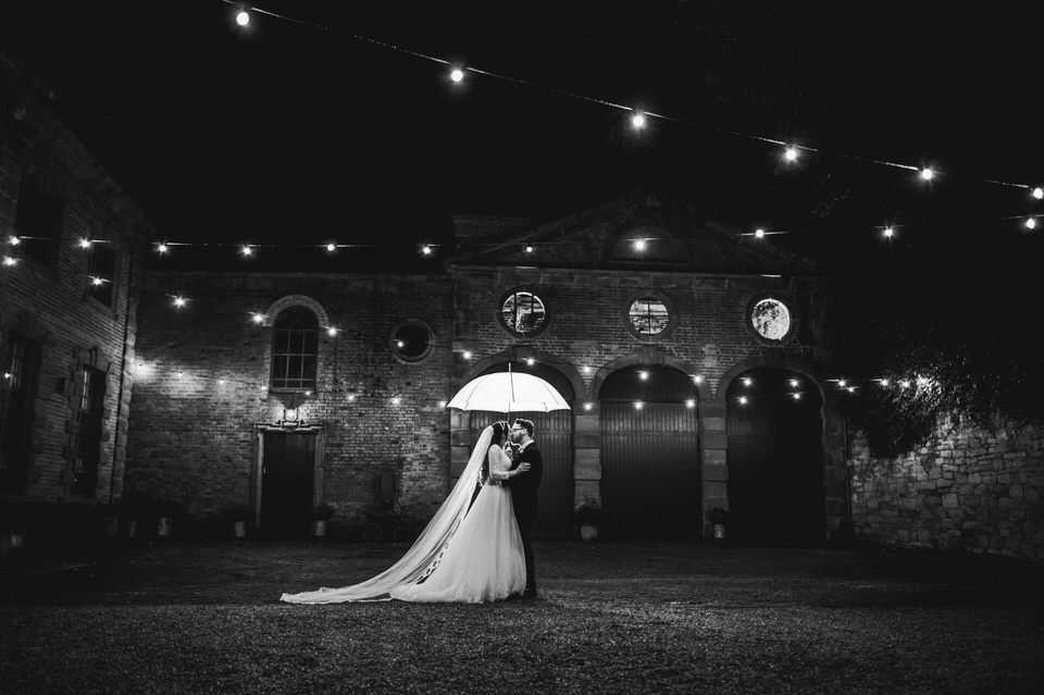 Bride & Groom outside Soughton hall wedding venue at night time, holding umbrella under fairy lights