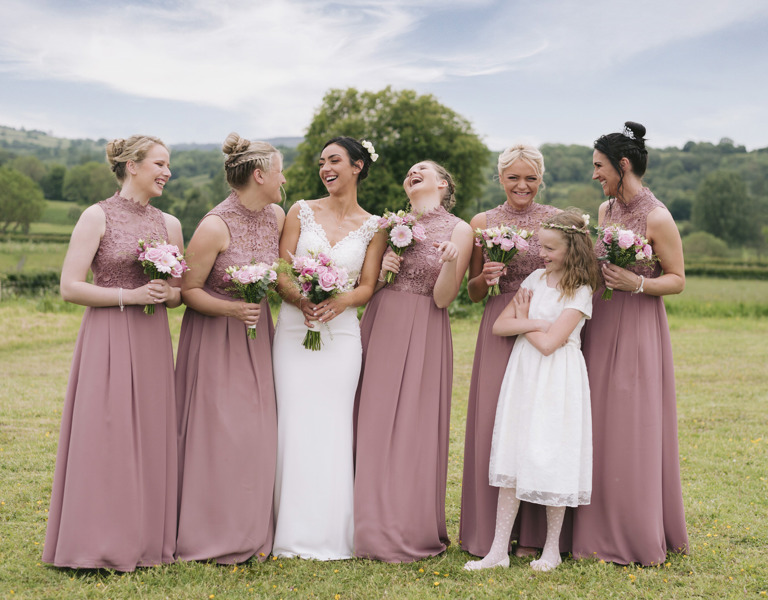 Bridesmaids laugh in pink dresses along with bride in North Wales field during wedding
